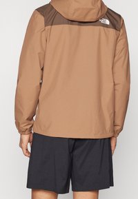 Veste imperméable marron avec un panneau d'épaule marron foncé, des poignets élastiques et une capuche. Accompagnée de shorts foncés.