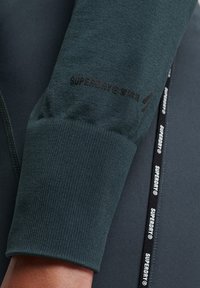 Prenda de color verde oscuro de manga larga con puños acanalados y un logotipo impreso en la manga. Incluye un cordón negro con el texto de la marca.