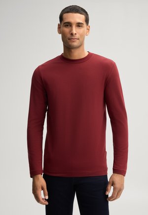 Man met kort donker haar, met een rood shirt met lange mouwen en een ronde hals, en donkere broek, staand tegen een effen lichte achtergrond.