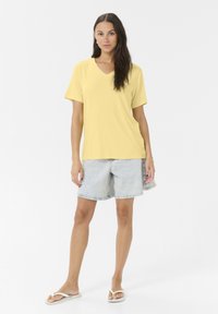 Gul V-udskæring t-shirt, korte ærmer, parret med lyseblå denim shorts, flade hvide sandaler, stående på en ensfarvet hvid baggrund.
