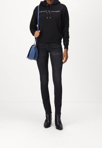 Schwarzer Kapuzenpullover mit weißem "Tommy Hilfiger"-Logo, dunkle Skinny-Jeans, schwarze Knöchel-Stiefel und eine blaue Handtasche mit Obergriff und Tragegurt.
