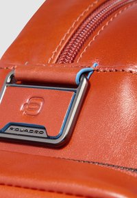 Detalle de mochila de cuero con un acabado en naranja brillante, un acento de herrajes metálicos plateados y un tirador de cremallera azul. Textura lisa con costuras visibles.