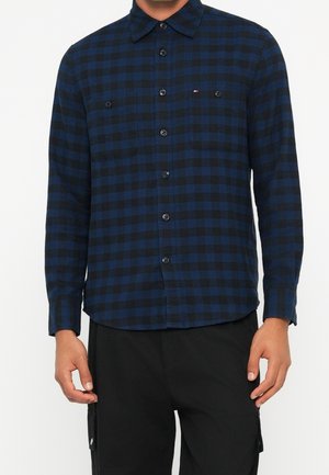 Homme portant une chemise à carreaux bleue et noire à manches longues avec deux poches poitrine et un pantalon noir.