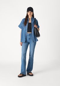 Camisa de mezclilla de botones, de manga corta; top negro corto; jeans de campana de color azul claro; gorra negra; bolso negro de hombro; chanclas negras.