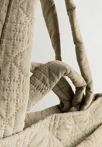 Gros plan sur un sac en tissu beige matelassé, mettant en valeur des poignées finement cousues entrelacées, sur un fond clair.