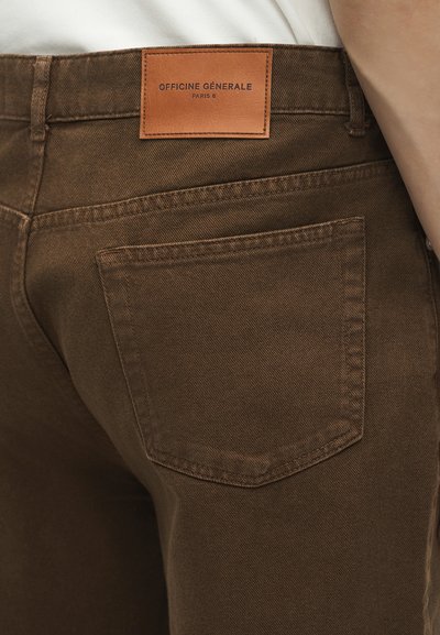 Κοντινή λήψη καφέ τζιν παντελονιού που δείχνει την πίσω τσέπη και δερμάτινο ταμπελάκι με την επωνυμία "Officine Générale Paris 6."