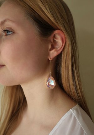 Gouden oorbel met een grote, facetgeslepen, druppelvormige kristal in tinten roze en paars, opgehangen aan een dunne, gebogen haak.