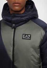 Mann trägt eine olivgrüne und marineblaue Steppjacke mit hohem Kragen und EA7 Emporio Armani Logo auf der Brust.
