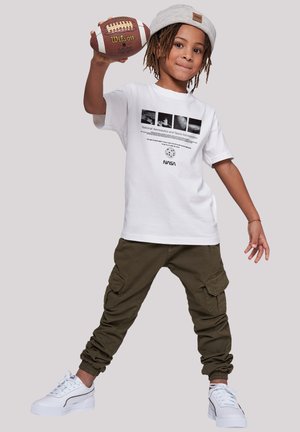 White cotton t-shirt with NASA print, olive green cargo pants with pockets, and white sneakers. The child holds a brown football.