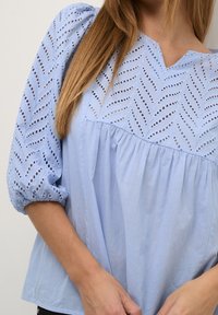 Ljusblå blus i ögonfäste med löv-mönster, puffärmar och en lös passform med en samlad bodice och V-ringning.