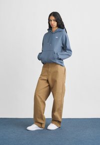 Μπλε φλις φούτερ με κορδόνια και μπροστινή τσέπη, συνδυασμένο με καφέ παντελόνι φαρδύ. Άσπρα sneakers συμπληρώνουν το σύνολο.