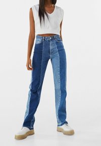 Blaue Jeans aus Denim mit kontrastierenden hellen und dunklen Bereichen, gerade geschnitten, hoher Bund und rohem Saum. Kombiniert mit einem weißen, kurzen Oberteil und klobigen Schuhen.