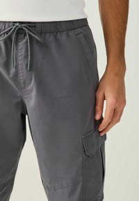 Homme portant un pantalon cargo gris foncé avec taille à cordon et poches latérales, main reposant près de la cuisse, fond clair.