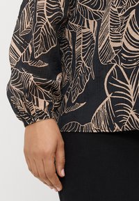 Blusa preta com um padrão de folhas em bege. Apresenta mangas bufantes com punhos elásticos e um tecido texturizado. Usada com calças escuras.
