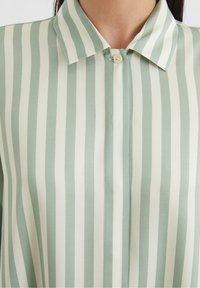Blusa a righe verdi e bianche con colletto classico, chiusura con bottoni e texture del tessuto liscia. Le righe verticali sono equidistanti.