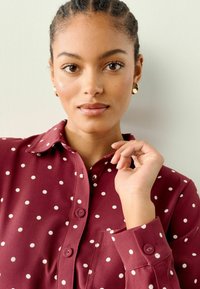 Burgundská košile s bílými puntíky, zapínání na knoflíky, skládací límec a manžety s nápletem, obsahující přední kapsu a hladkou texturu.