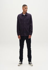 Camisa de botones de color púrpura oscuro con dos bolsillos en el pecho, combinada con jeans oscuros y zapatillas negras. Modelo de pie contra un fondo neutro.