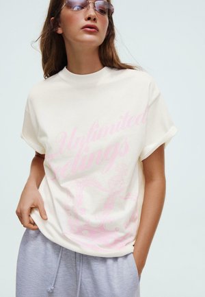 Nő napszemüvegben, fehér pólóban, amelyen rózsaszín "Unlimited Feelings" felirat olvasható, és világosszürke zsinóros nadrágban, egyszínű háttér előtt pózol.