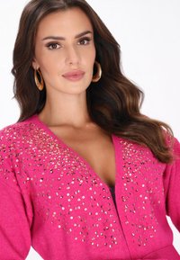 Hosszú barna hajú nő nagy arany karika fülbevalókkal és fényes flitterekkel díszített élénk rózsaszín pulóverben.