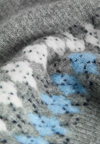 Close-up van zachte gebreide stof met een geometrisch patroon in grijs, wit en blauwe kleuren, waarbij textuurvezels en stikseldetails te zien zijn.