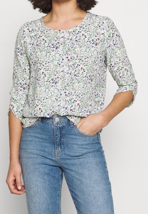 Kobieta w białej bluzce z małym wielokolorowym kwiatowym wzorem i podwiniętymi rękawami, w połączeniu z jasnoniebieskimi, wysokim stanem jeansami.