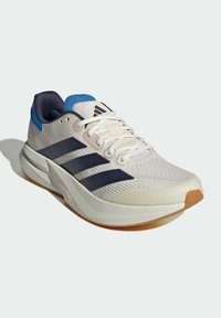 Chaussure de course Adidas beige clair avec des bandes bleu marine, un patch sur le talon bleu, des lacets blancs et une épaisse semelle blanche avec un bout de couleur gomme.