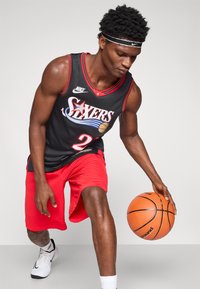 Μαύρη μπλούζα μπάσκετ με κόκκινες λεπτομέρειες και αριθμό παίκτη 23, συνδυασμένη με κόκκινες βερμούδες και λευκά αθλητικά παπούτσια, κρατώντας μια μπάλα μπάσκετ.