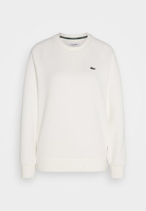 Valkoinen puuvillaneule, jossa on ribbireunat hihoissa ja helmassa. Ominaisuuksiin kuuluu pyöreä pääntie ja vihreä Lacoste-logo kauluksessa. Klassinen malli.