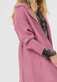 Femme aux longs cheveux blonds portant un long cardigan rose sur un chemisier sombre à fleurs, coupée au niveau du milieu du visage, debout devant un fond clair et uni.