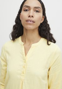 Chemise jaune à manches longues en tissu léger. Dotée d'un col rond, d'une patte de boutonnage et d'une texture subtile.