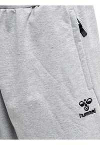 Grijze sweatpants van zachte stof, met een zwart logo en een met rits afgesloten zak aan de zijkant. De textuur lijkt een beetje pluizig.