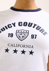 Vit bomulls t-shirt med mörkblå halsringning. Har "Juicy Couture", en sköldlogotyp och fem stjärnor tryckta i fet marinblå. 1997 års kaliforniska tema.