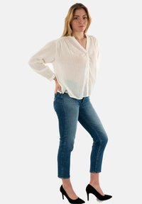 Blusa blanca ligera con botones en la parte delantera y sutiles rayas verticales, combinada con jeans ajustados azules y tacones de aguja negros.