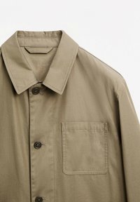 Khaki-jack met een knoopsluiting aan de voorkant, een puntige kraag, een enkele borstzak en een gladde textuur. Opmerkelijke donkere knopen en stikdetails.