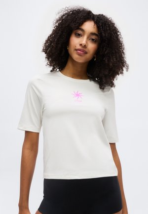 Femeie cu păr creț purtând un tricou alb cu mânecă scurtă, cu un mic desen roz de soare pe piept, și pantaloni negri.