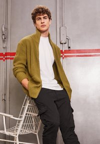 Olivgrön rippad cardigan över en vit t-shirt, tillsammans med svarta cargobyxor; modell står bredvid en vit metallstol.