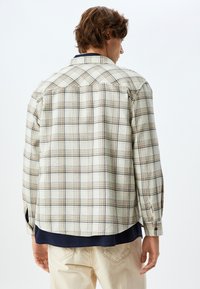 Camisa de cuadros de manga larga en beige claro con líneas azules y marrones. Tiene puños con botón y un dobladillo curvado, realizada en una tela texturizada.