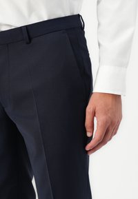 Pantaloni tailored de culoare bleumarin, dintr-un material neted, cu fața plată, buzunare laterale și talie adaptată cu bucle pentru curea. Manșeta unei cămăși albe este vizibilă.