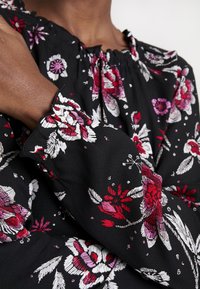 Blouse noire à motif floral avec des fleurs rouges et roses, encolure froncée, et manches longues avec poignets élastiques. Fabriquée en tissu léger et texturé.