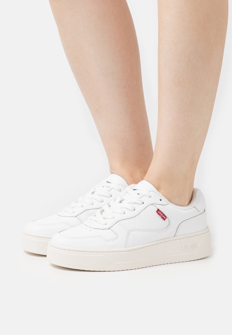 Zapatillas blancas con parte superior de cuero texturizado, cordones planos, un collar acolchado y una suela de goma beige clara con un detalle de logo en el lado.