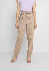 Kvinna iklädd hög midja beiga rutiga byxor med knytbälte, tillsammans med en lavendelfärgad ärmlös topp och lavendelfärgade sandaler med klack och remmar.