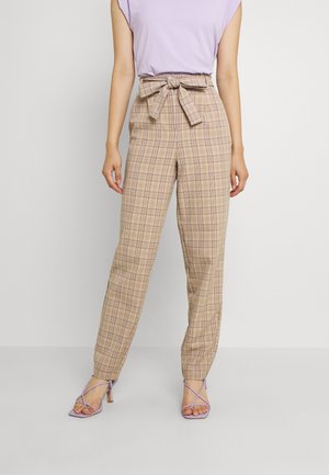 Kvinna iklädd hög midja beiga rutiga byxor med knytbälte, tillsammans med en lavendelfärgad ärmlös topp och lavendelfärgade sandaler med klack och remmar.