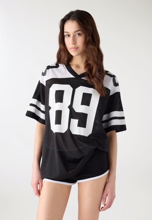 Giovane donna con capelli lunghi che indossa una maglia sportiva oversize bianca e nera con il numero 89 e pantaloncini atletici neri su sfondo semplice.
