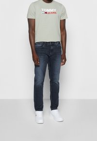 Ljusgrön bomullströja med "TOMMY JEANS" tryckt i vitt och rött, kombinerad med mörkblå denimjeans och vita sneakers.