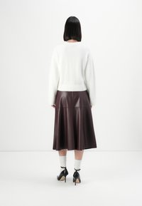 Vrouw met recht, zwart haar draagt een witte pluizige trui, een donkerbruine leren midi-rok, witte sokken en zwarte hoge hakken, staand tegen een witte muur.
