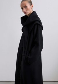 Abrigo largo negro con cuello sobredimensionado, mangas anchas y bolsillos delanteros. Confeccionado con una tela texturizada, presenta una silueta recta.