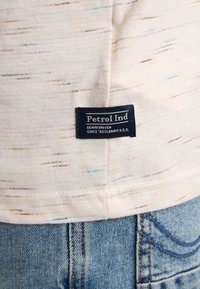 Lichtbeige stof met kleurrijke stippen; heeft een blauw label met de tekst "Petrol Ind" in witte letters; gedragen met een spijkerbroek.