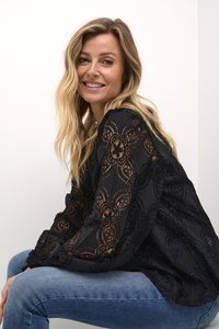 Črna čipkasta bluzica z rožnatimi vzorci, prosojno teksturo in dolgimi rokavi, v kombinaciji z modrimi kavbojkami. Oblikovanje prikazuje zapleteno detajliranje.