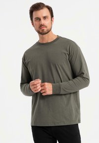 Mann mit leichtem Bart, der ein olivgrünes Langarmshirt und schwarze Hosen trägt, steht vor einem schlichten weißen Hintergrund.