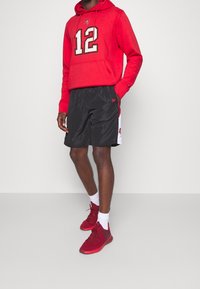 Roter Kapuzenpullover mit der Nummer 12, schwarze Sportshorts mit weißen und roten Akzenten sowie roten Sportschuhen. Lässiges Sportbekleidungsensemble.